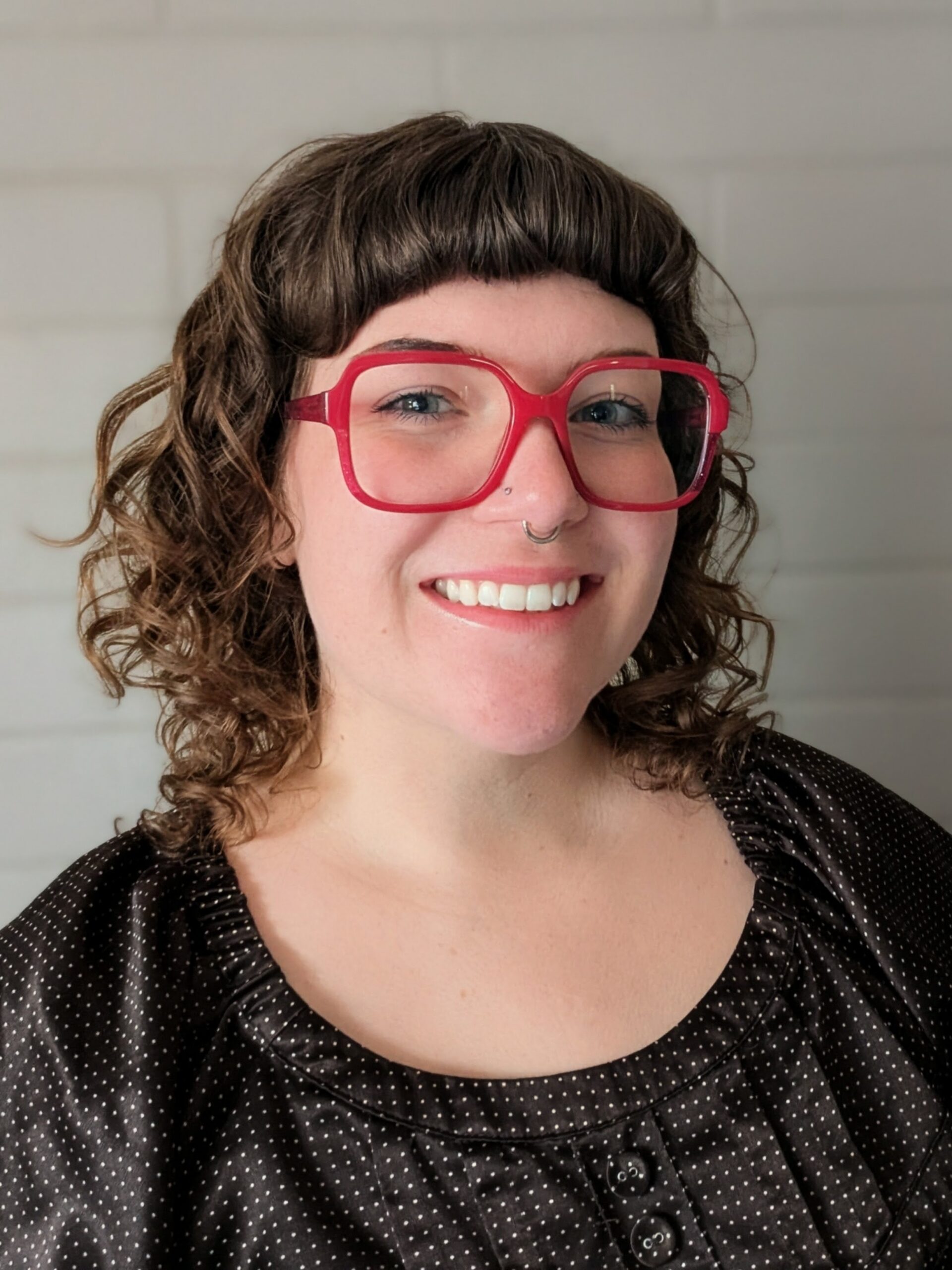 Close-up of a smiling woman with bright red oversized glasses, dark curly hair with bangs, wearing a black polka-dot blouse indoors.