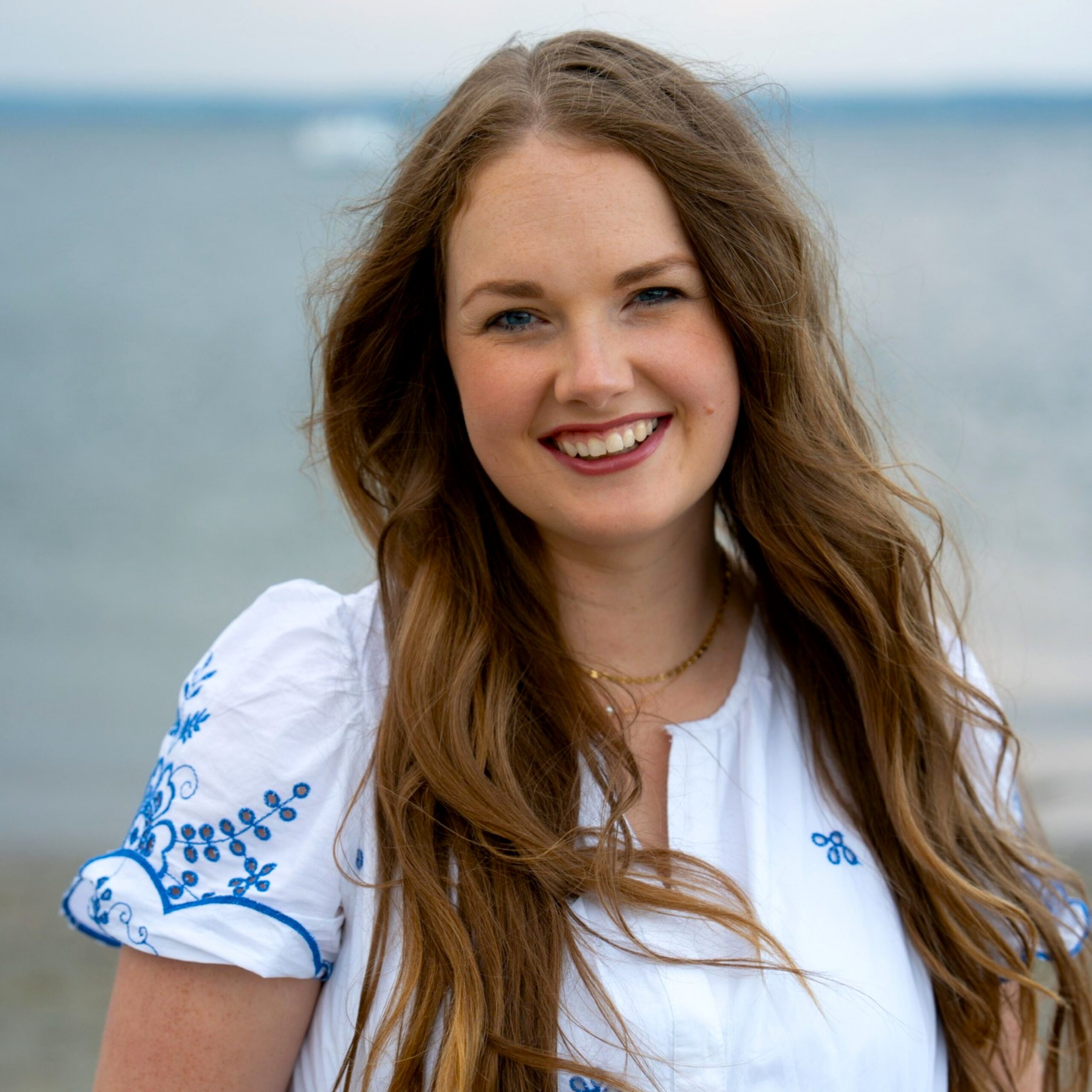 Madison Christensen, LMFT | The Evolution Group Smiling woman with long brown hair at the beach, wearing a white blouse with blue embroidery on the sleeves and chest.