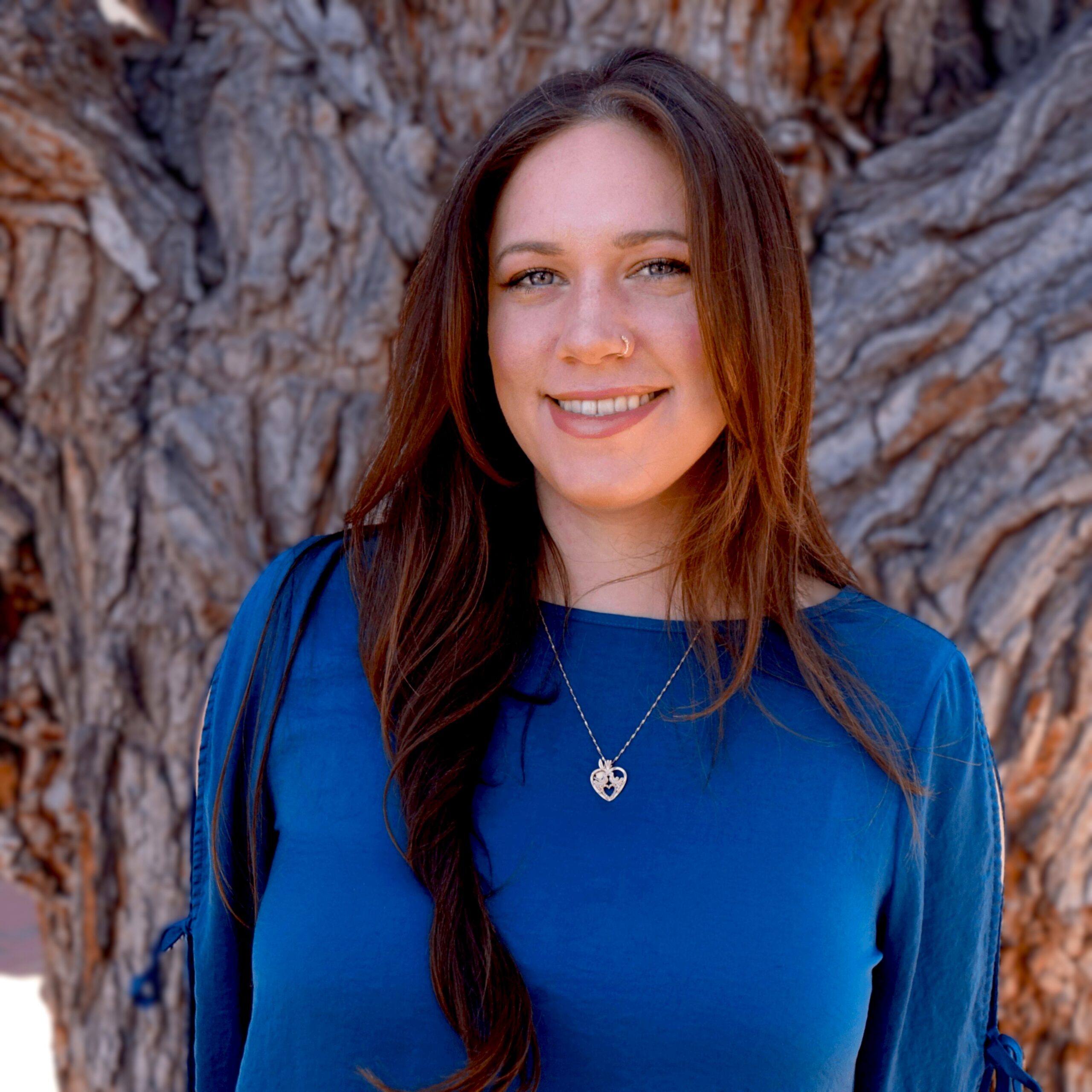 Mallory Hallstead, LMSW | The Evolution Group Portrait of a smiling woman with long brown hair wearing a blue top, standing in front of a textured tree trunk backdrop, with a heart-shaped necklace visible.